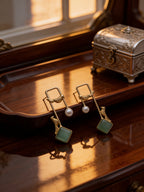 Pair of earrings with green / emerald square stones and pearl-like centers on a wooden surface.