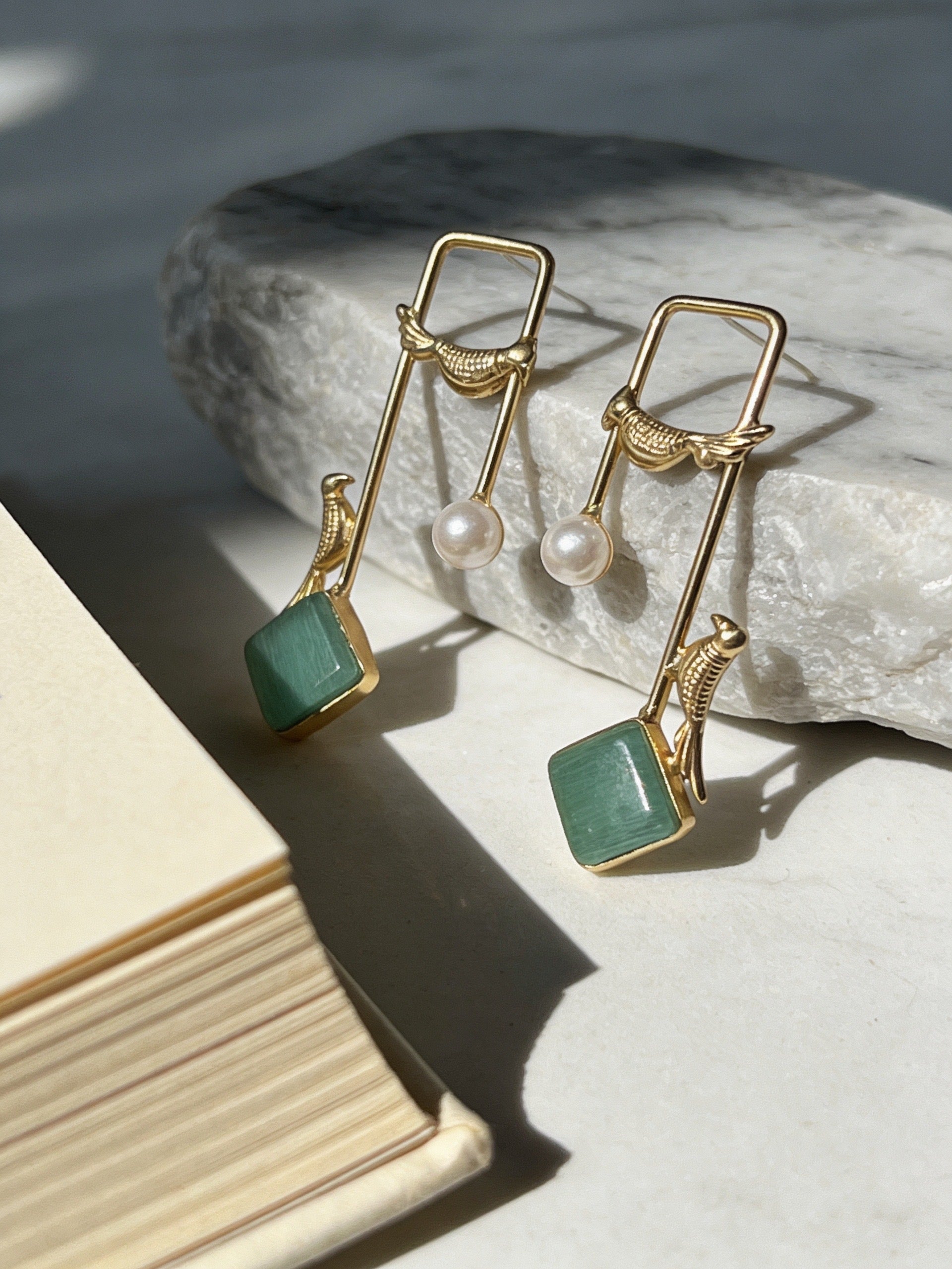 Gold earrings with green square stones and pearls on a stone surface